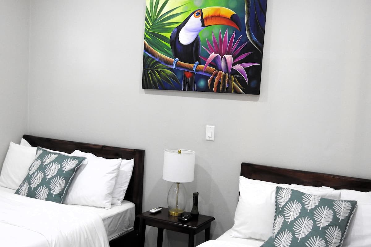 Quarto de hotel com duas camas, roupa de cama branca, almofadas com padrão de folhas verdes, mesa de cabeceira com luminária e um quadro de um tucano na parede.