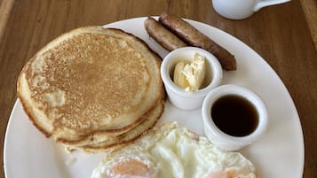Un piatto con pancake, due fette di pancetta, un uovo fritto, una piccola ciotola di burro e una tazza di caffè su un tavolo di legno.