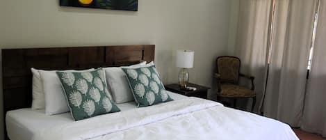 Una habitación de hotel con una cama grande, una mesita de noche con lámpara, una silla y un cuadro de tucanes en la pared.
