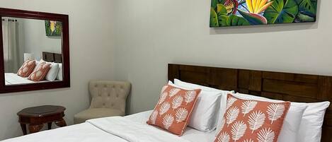 A hotel room with a bed, a chair, a round table, a mirror, and a painting of a bird and leaves on the wall.