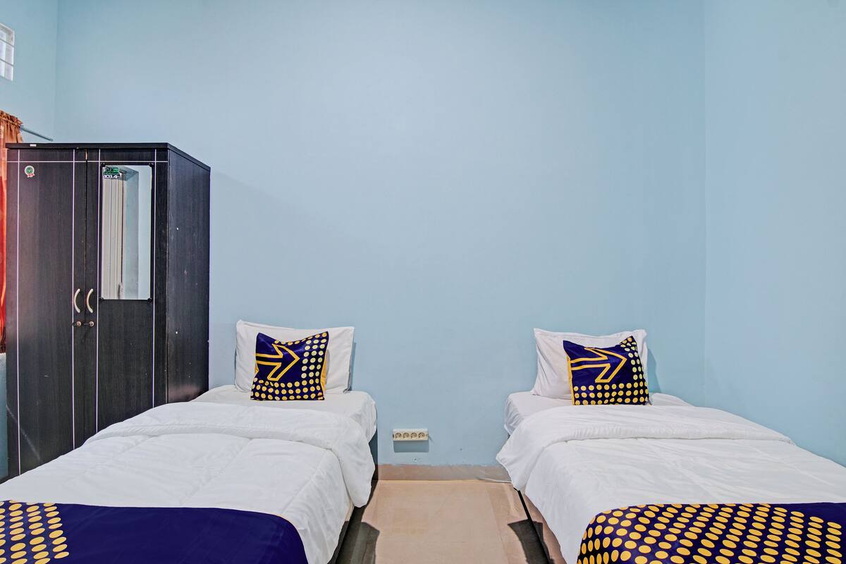 Una habitación con dos camas, cada una con un colchón blanco y almohadas estampadas en azul y amarillo. Se observa un armario de color oscuro en el lado izquierdo de la habitación.