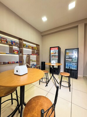 A small dining area with a round table, two stools, a toaster, shelves with snacks, and two beverage dispensers. - Pousada Leda Rafael (Joao Camara)