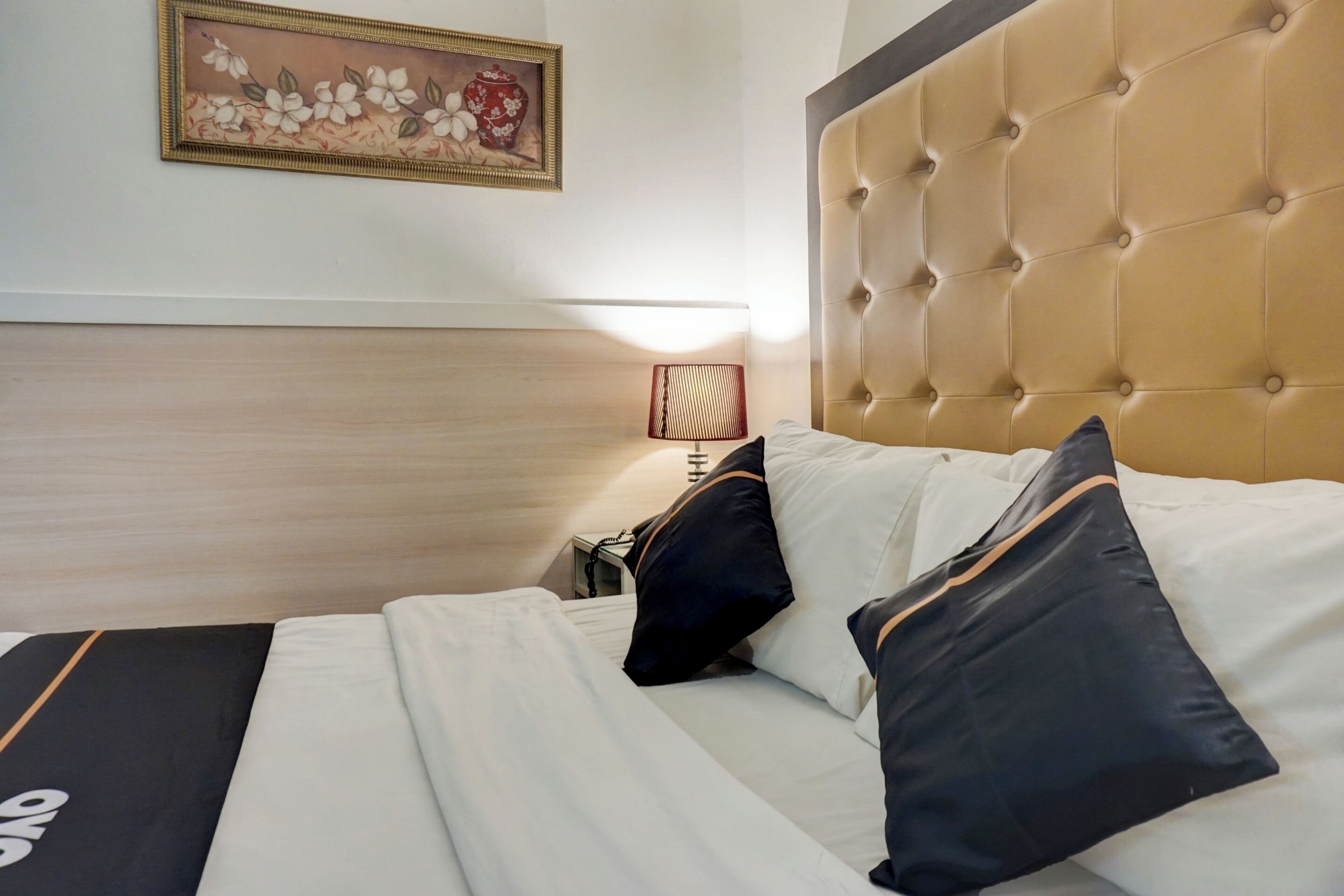 A hotel room with a tufted headboard, a bed with white and black bedding, a framed floral artwork on the wall, and a bedside lamp.