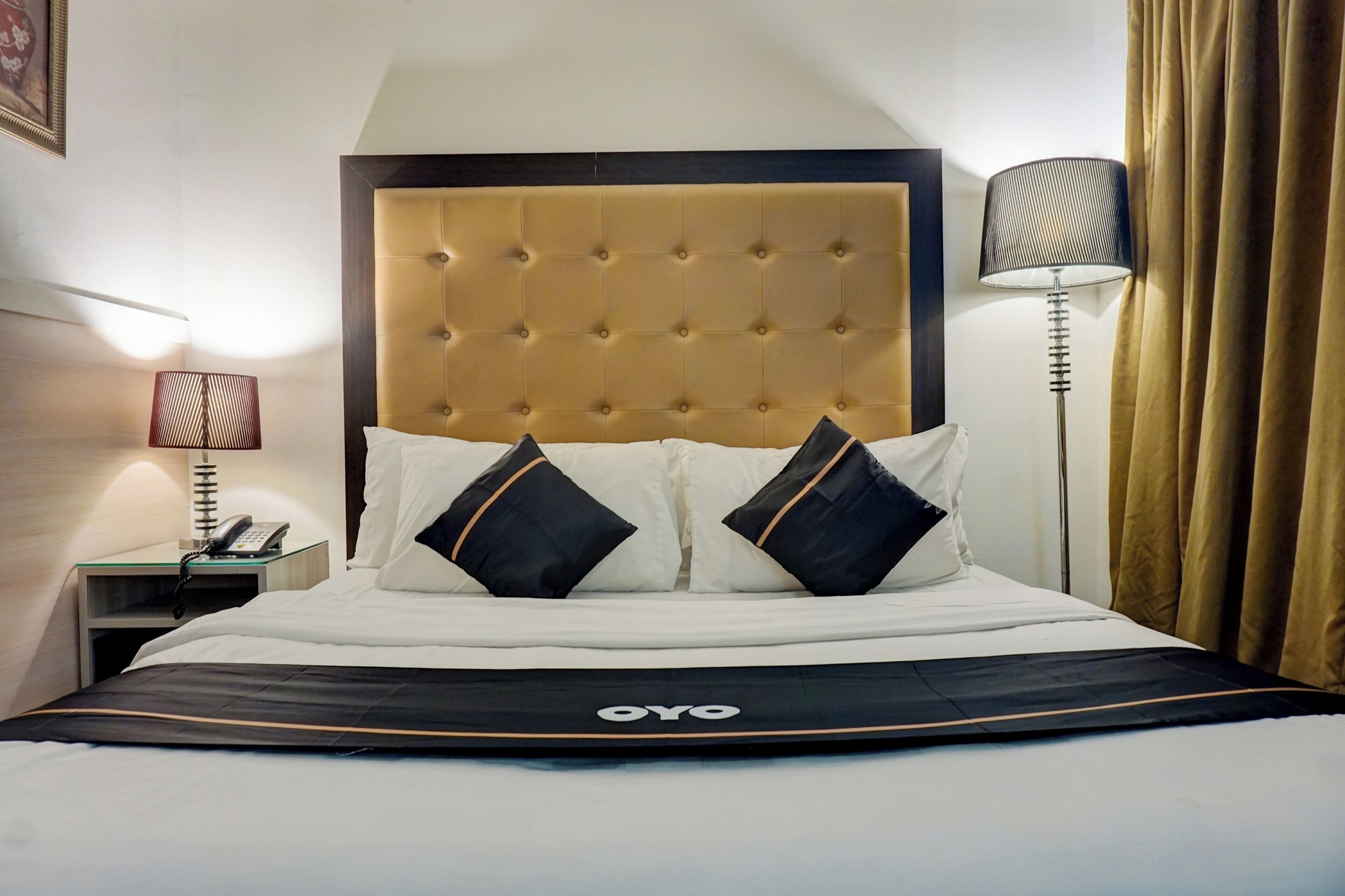 Una habitación de hotel con un cabecero capitonado, una cama con ropa de cama blanco y negro, una lámpara de noche y un teléfono sobre una mesa de vidrio.