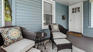 Une véranda avec deux fauteuils en osier et une petite table, une porte blanche et une fenêtre avec des stores.