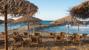 Una playa con sillones de madera y sombrillas de paja, frente a un mar azul y despejado.