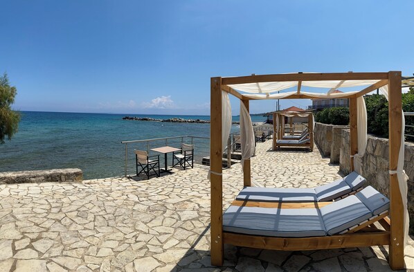 A wooden outdoor seating area with a canopy, stone flooring, and a view of the sea. - Amaltheia Beach Front Houses (Zakynthos)
