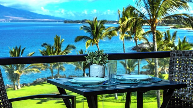 Una mesa de comedor con platos y cubiertos, un vaso de agua y una planta pequeña en un balcón con vistas a una playa con palmeras y el océano.