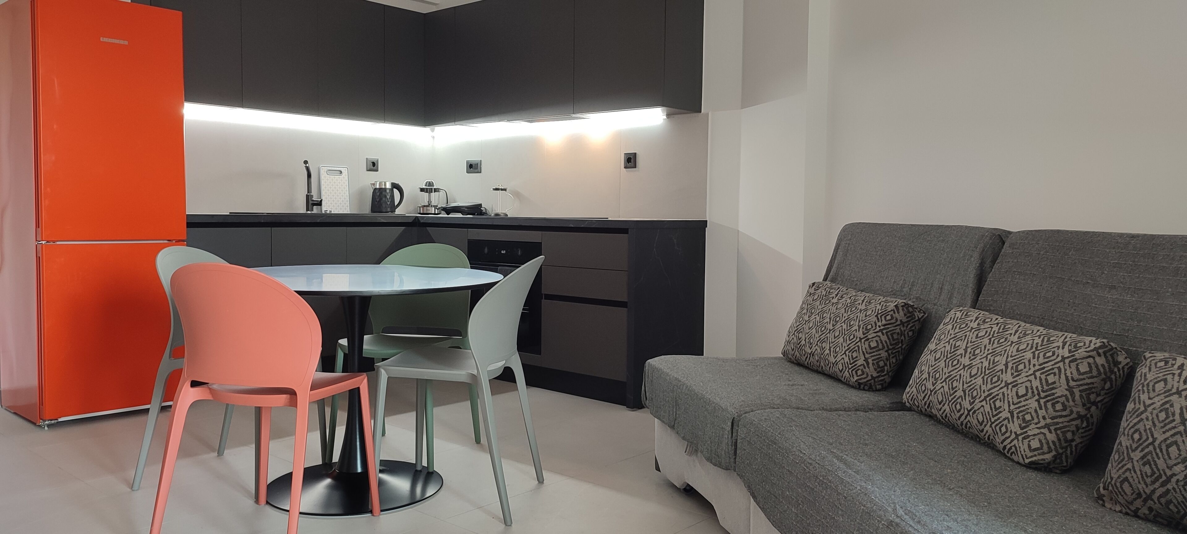 Une cuisine compacte dotée d’appareils électroménagers modernes, une table à manger avec des chaises et un canapé gris agrémenté de coussins à motifs.