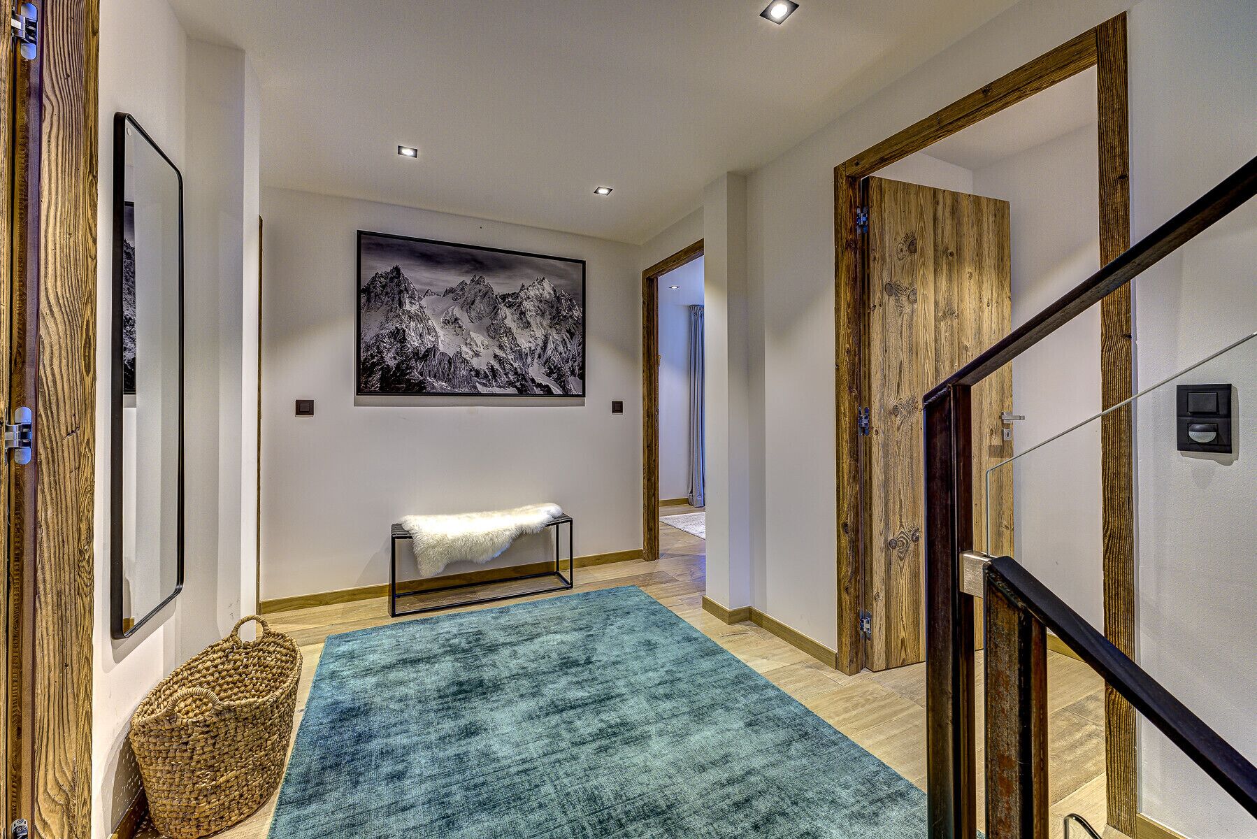 Un couloir avec une porte en bois, un tableau encadré représentant des montagnes, un banc recouvert d’une couverture blanche et un tapis bleu.