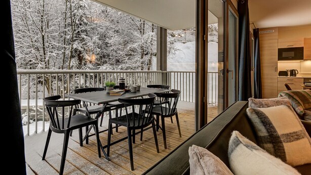 Un balcon avec une table et des chaises, un canapé et un coin cuisine visible à travers une porte vitrée.