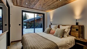 Une chambre avec un plafond en bois, un grand lit recouvert de plusieurs oreillers, une table de chevet et une vue sur un paysage enneigé, visible à travers une porte coulissante.