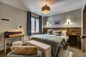 A modern bedroom with a wooden headboard, a large bed with multiple pillows, a desk with a lamp, and a view of a snowy landscape through the window. - Duplex Apartment with Balcony &amp;amp; Mountain Views (Chamonix-Mont-Blanc)