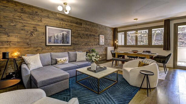 Un salon moderne avec un mur en bois, un canapé d’angle, une table basse et un coin repas baigné de lumière naturelle grâce à de grandes fenêtres.