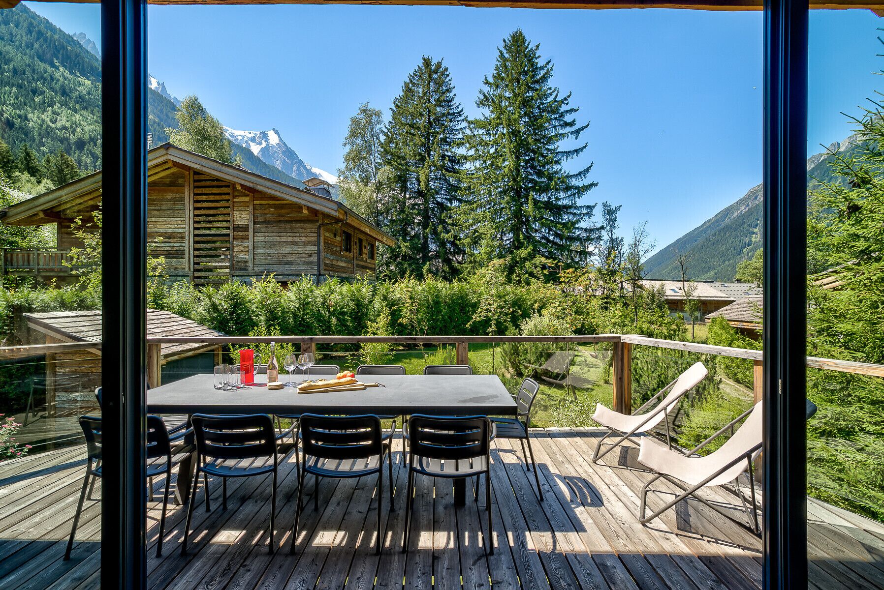 Una terraza de madera con una mesa y sillas, con vista a una cabaña en la montaña y un bosque.
