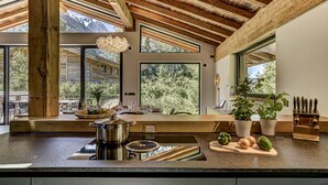 A modern kitchen with a large island, wooden beams, and large windows offering views of the outdoors. - Serviced Luxury Chalet with Pool & Spa (Chamonix-Mont-Blanc)
