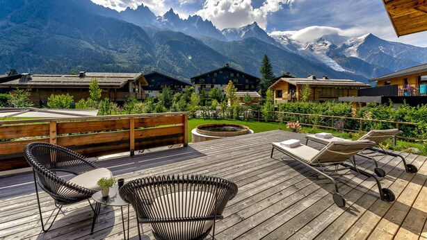 Eine Holzterrasse mit Liegestühlen und einem Whirlpool, mit Blick auf ein Gebirge mit Chalets.