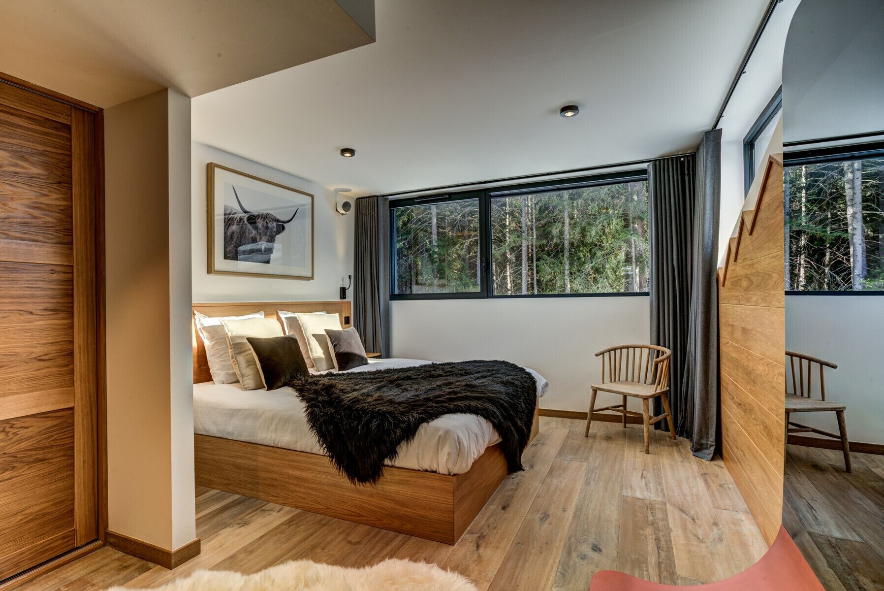 Un dormitorio moderno con una cama grande, cabecera de madera, un cuadro enmarcado, una ventana con vista a árboles y una silla.