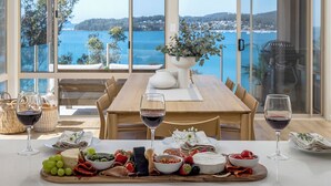 Una mesa preparada para una comida con vista al mar, que incluye una mesa de madera, platos, vasos y diversas opciones de comida.