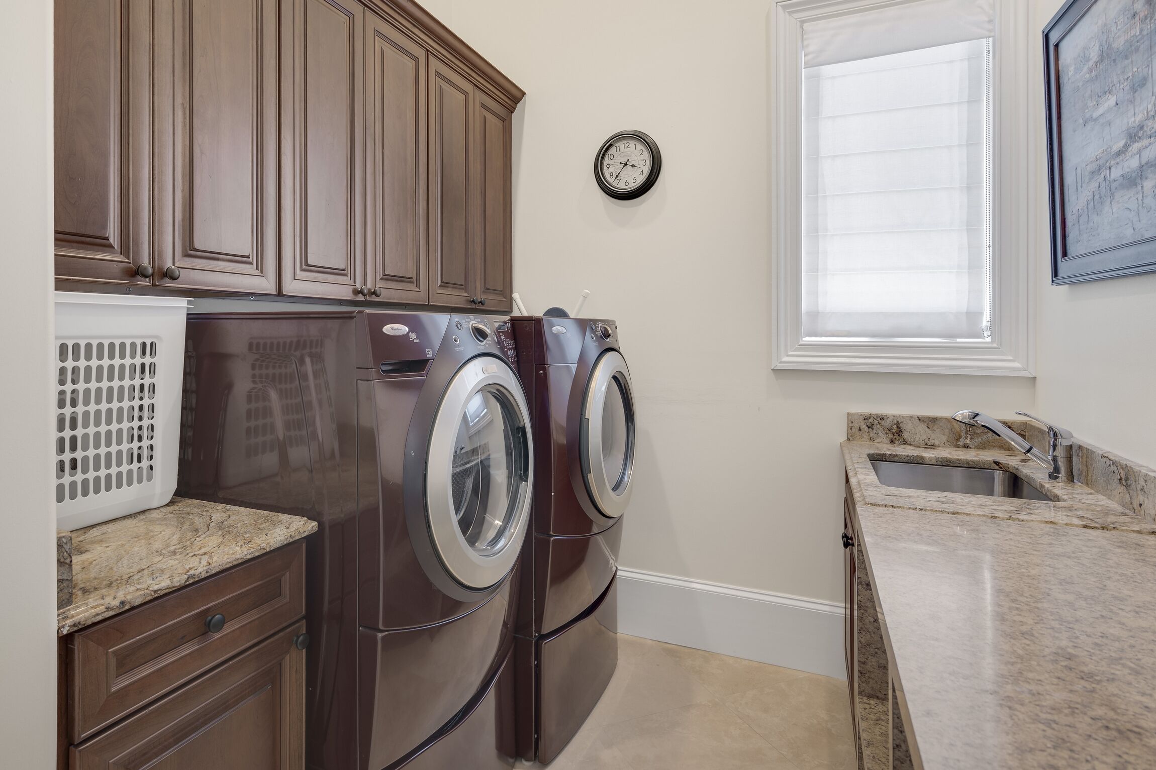 A laundry area with two front-loading washers, a drying rack, a clock, and a window with blinds.