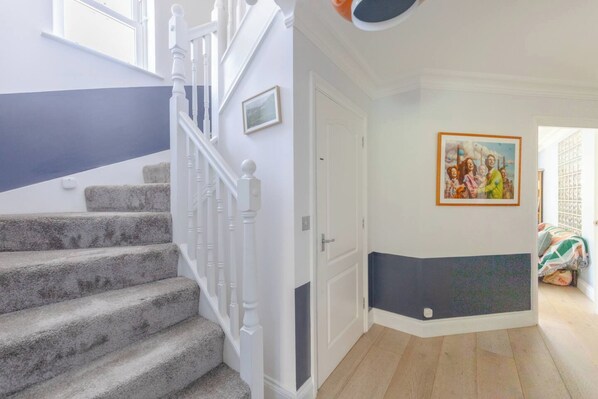 Un escalier avec des balustrades blanches, une porte dont la moitié inférieure est de couleur foncée et un tableau encadré accroché au mur.