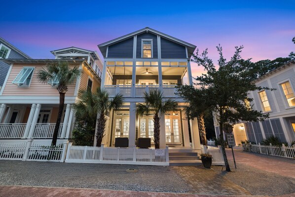 A two-story house with a balcony, palm trees, and a white picket fence.