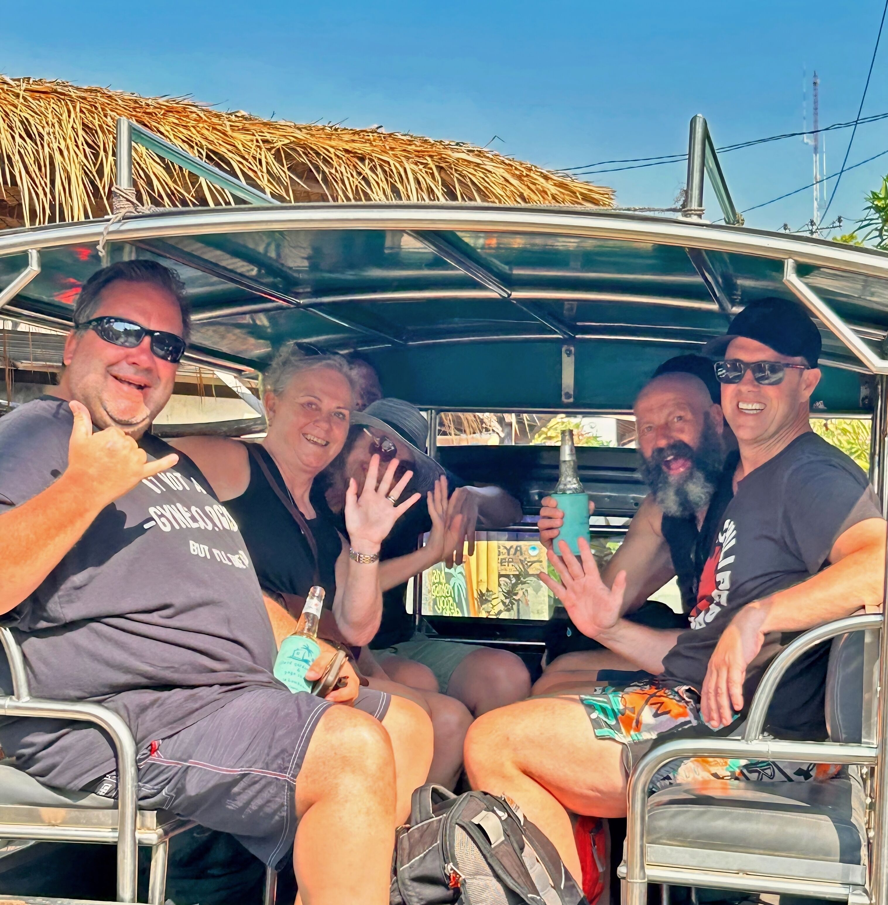 A group of people seated in a vehicle with a thatched roof, a metal frame, and a table with a bottle and a cup.