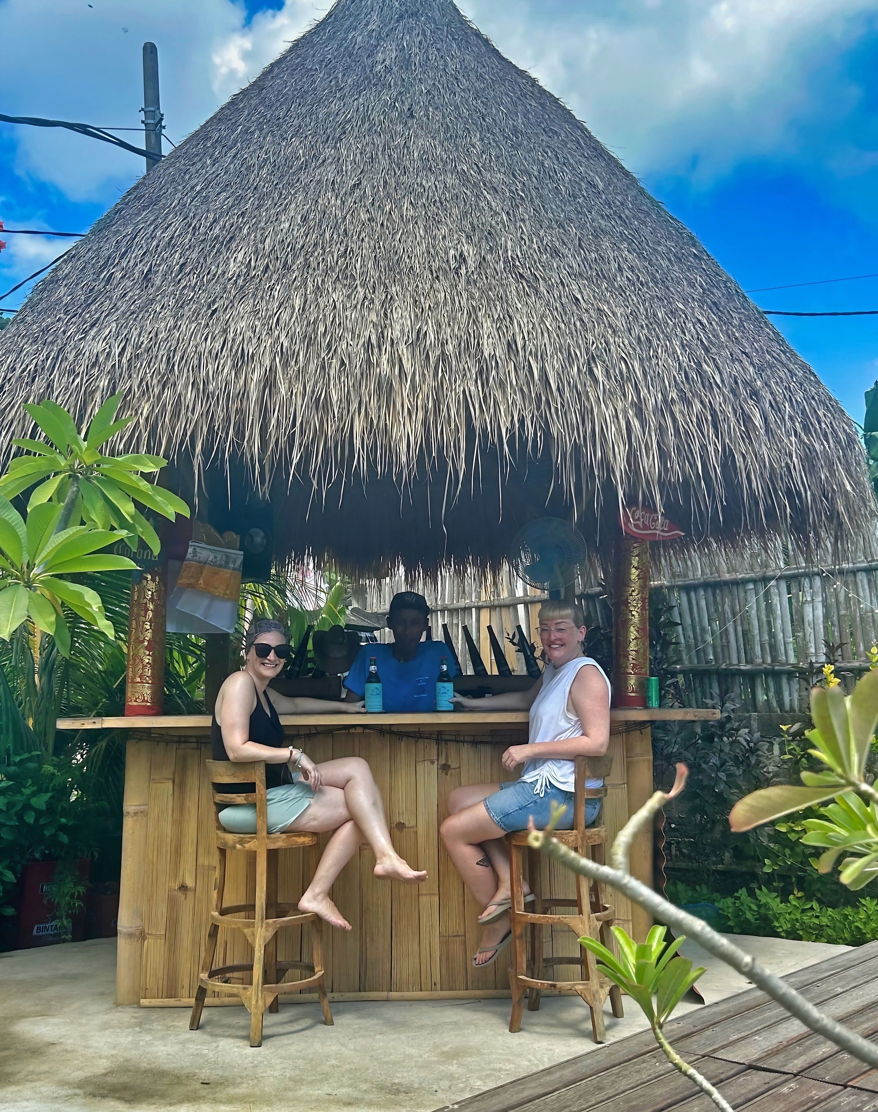 A thatched-roof bar with wooden stools, surrounded by greenery.