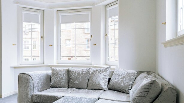 A corner room with a grey sofa, three windows, and a white wall. - Snug & Serene 2BD Flat - Roseburn (Edinburgh)