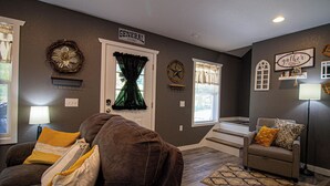Een woonkamer met een bruine bank, een grijze zithoek en een vloerkleed met dessin. Er is een deur met een bord erboven, een wandklok en een raam met gordijnen.