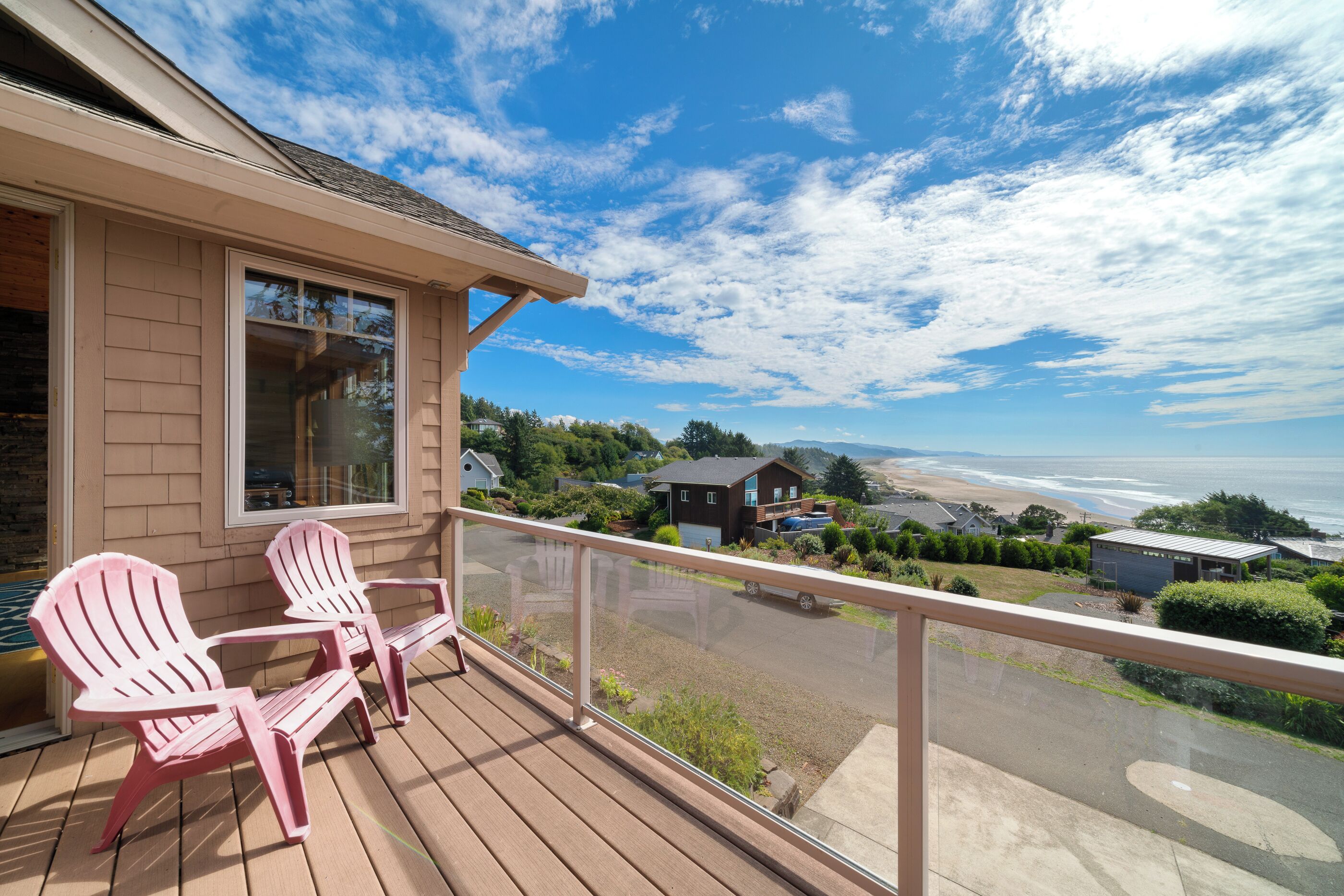 A wooden deck with two Adirondack chairs, a glass balustrade, and a view of a beach and ocean.