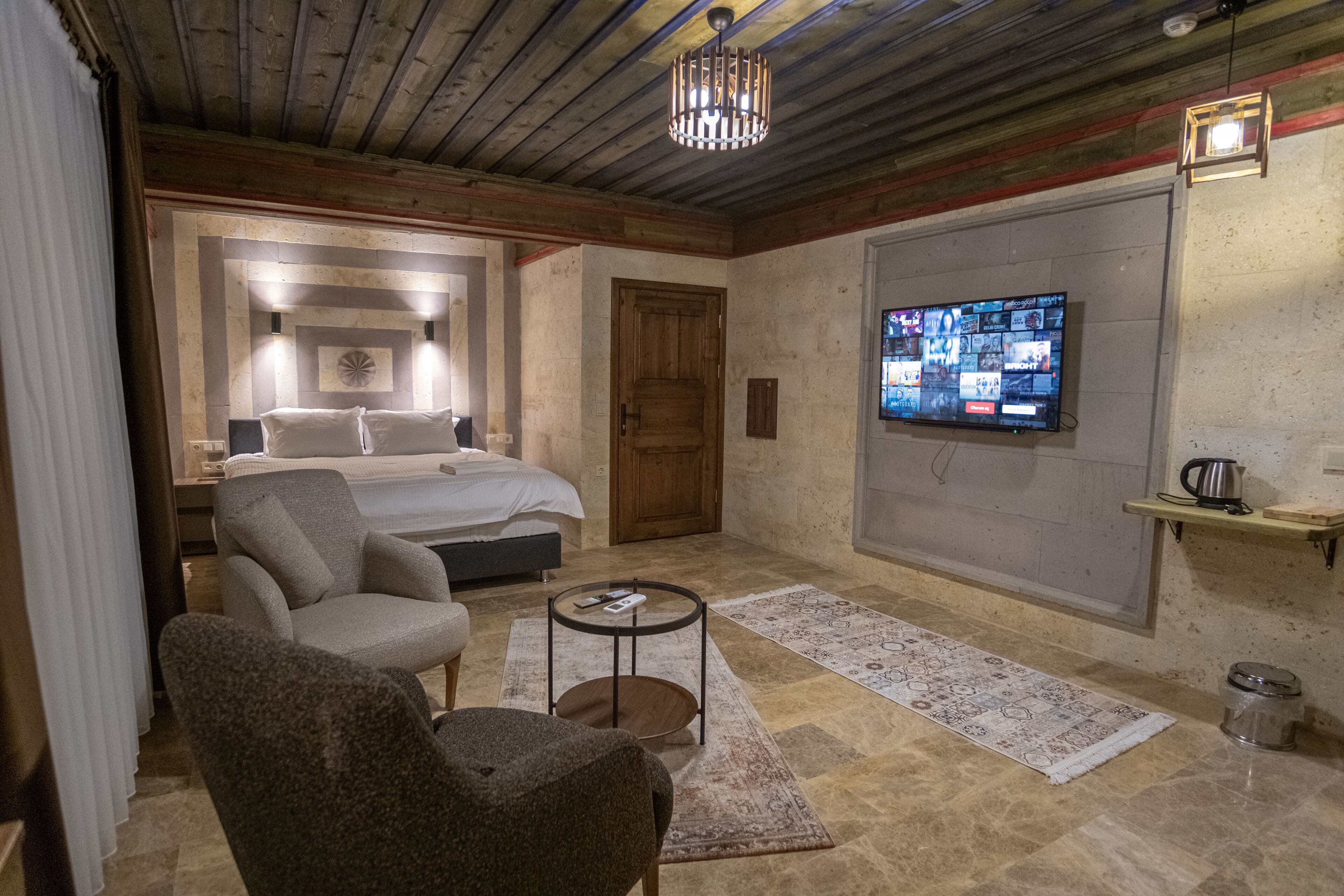 A cozy room with a bed, a chair, a small table, a TV mounted on the wall, and a wooden ceiling.