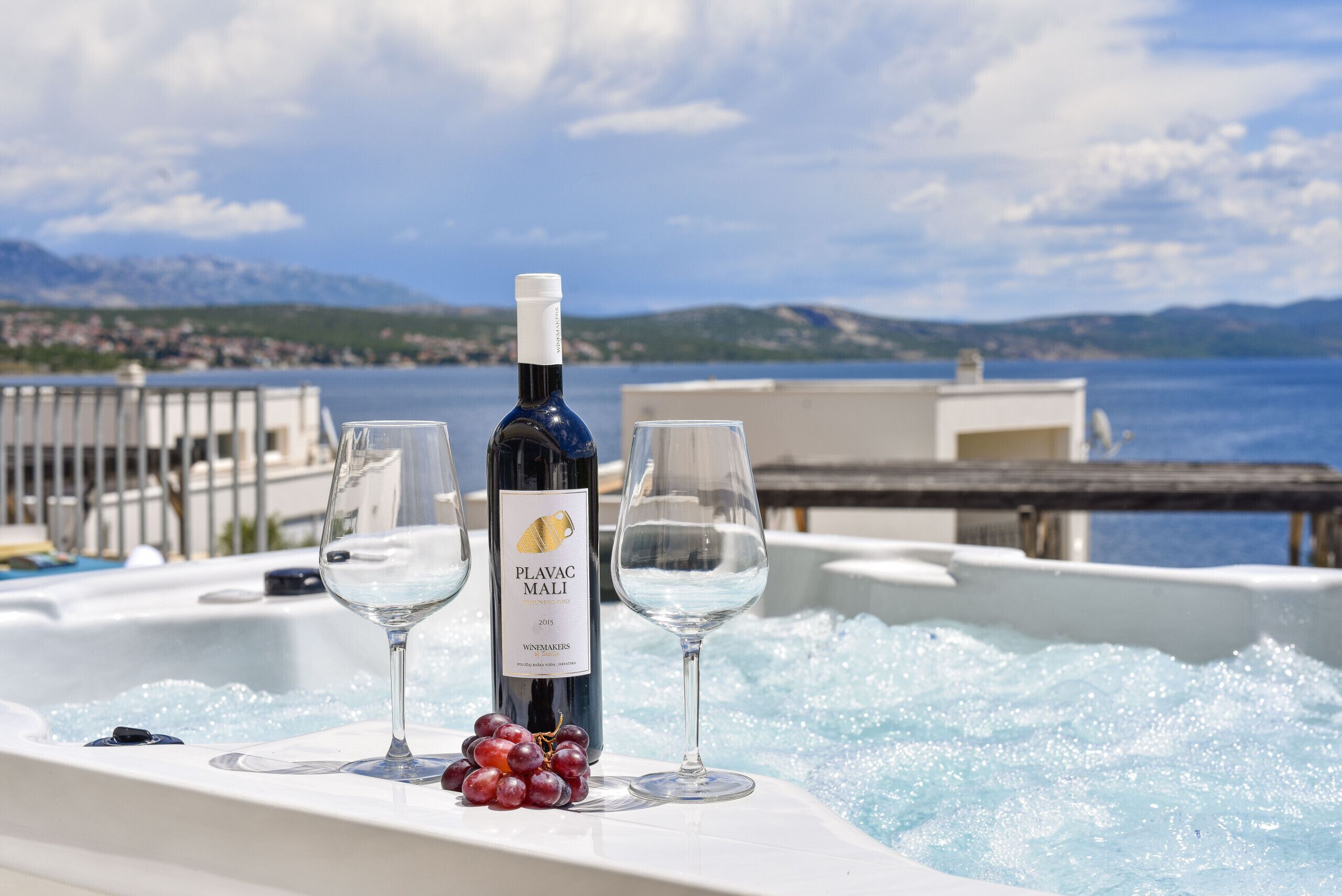 A bottle of Plavac Mali wine, two glasses, and grapes on a table with a view of the sea and mountains.