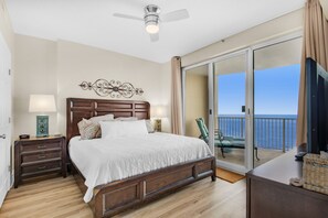 A bedroom with a large bed, a ceiling fan, a nightstand with a lamp, and a view of the ocean through a sliding glass door.