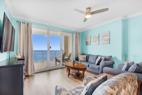 A spacious living room with a balcony, a ceiling fan, and a large window offering a view of the sea.