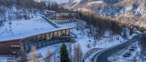 Un edificio moderno con fachada de vidrio, rodeado de montañas nevadas y una carretera sinuosa.