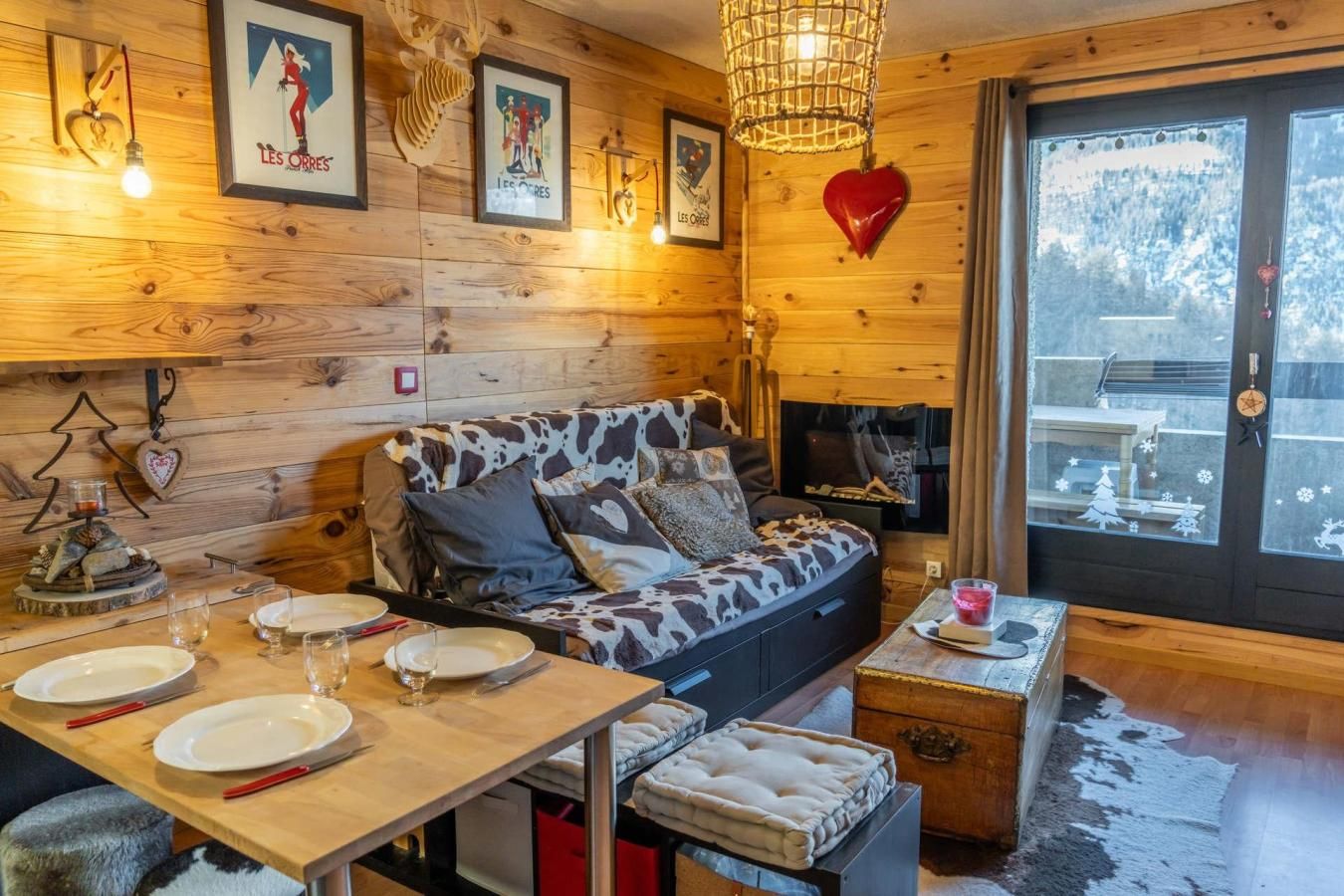 A cozy cabin interior with a wooden wall, a sofa with a cowhide pattern, a dining table set for two, and a fireplace.