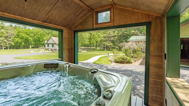 A wooden spa area with a hot tub and a view of a green house and trees. - Lakefront Oasis w/ Hot Tub, Pool, Courts & 2 Homes (Oakland)