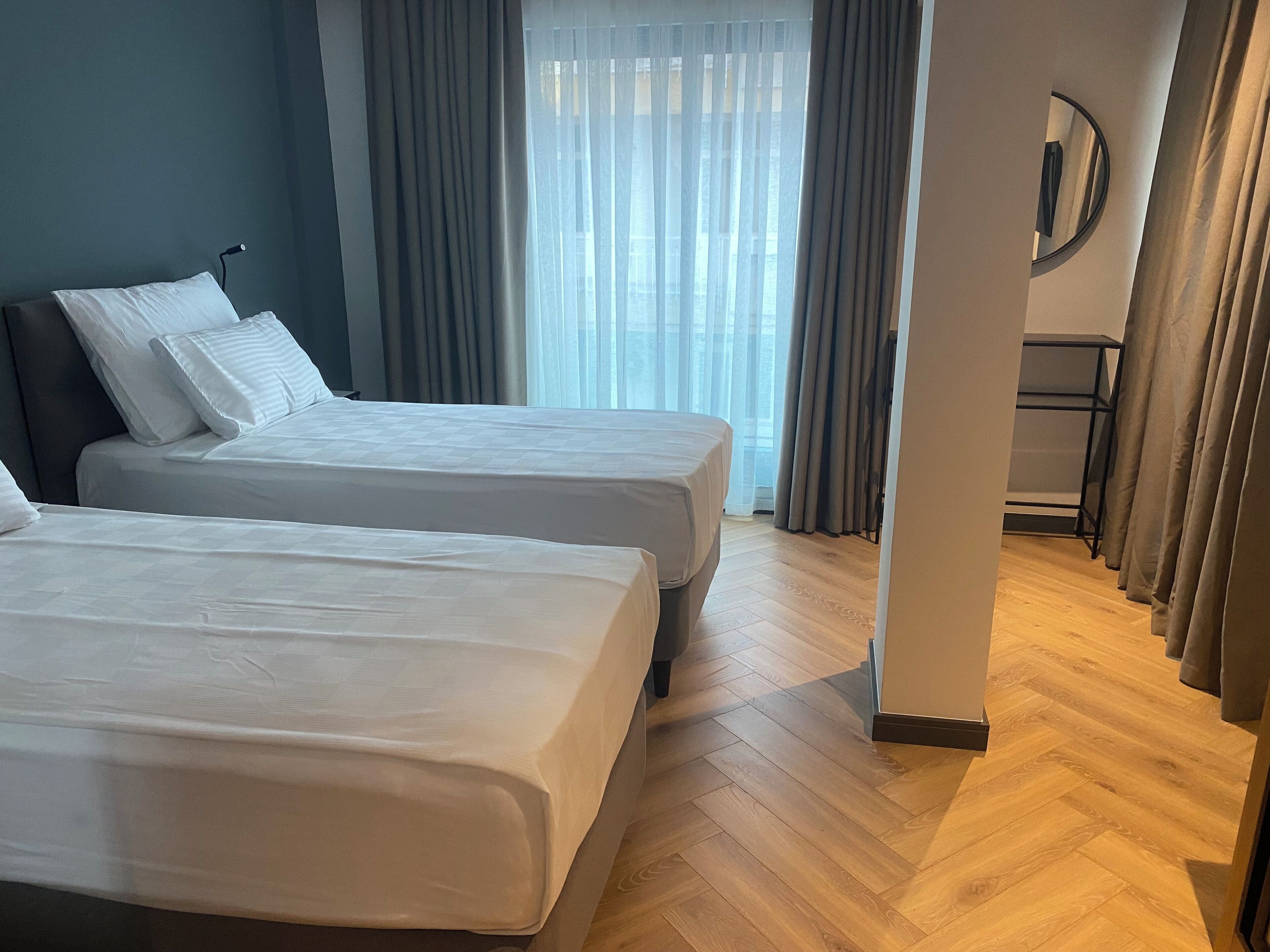Ein Hotelzimmer mit zwei Betten, Holzboden, Blick auf die Stadt durch transparente Vorhänge und einem kleinen Tisch mit Spiegel.