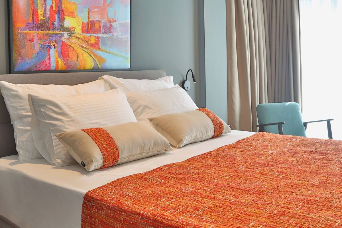 Une chambre d’hôtel avec un grand lit, un tableau au mur, une chaise et une fenêtre avec des rideaux.