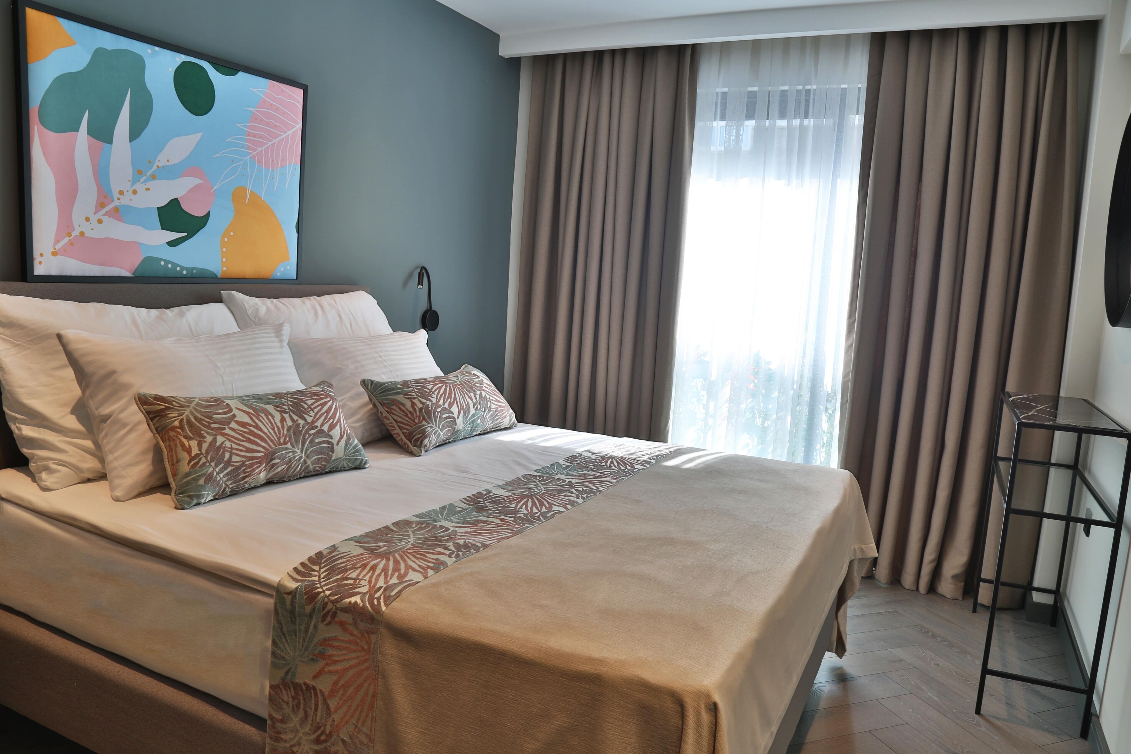 A neatly made bed with a floral bedspread, a large abstract painting on the wall, and a view of the outdoors through sheer curtains.