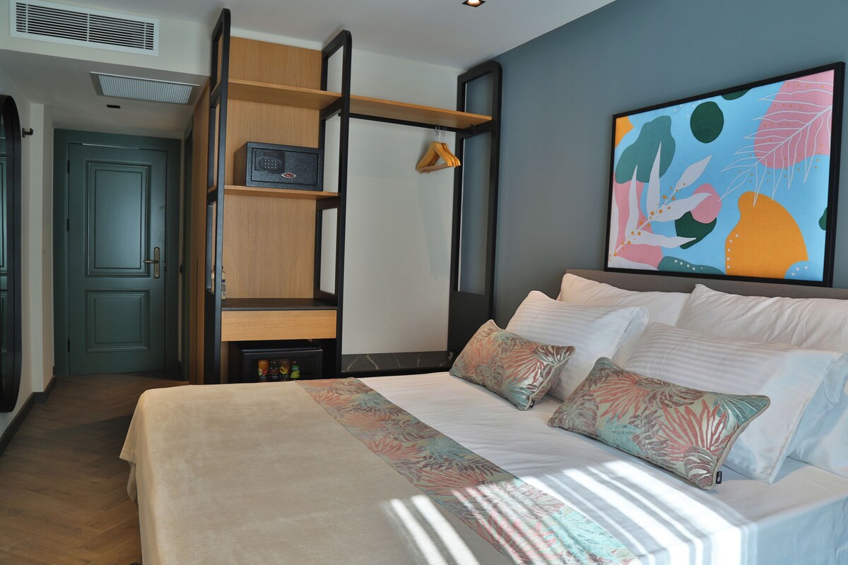 A modern hotel room with a bed, a wooden shelf unit, a painting, and a door.