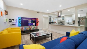 A modern living room with a sofa, coffee table, and a TV displaying streaming services. - 🕹️🎬🏎️👾Gamers Getaway: Arcade ,Theater, Racing, And More! (Fullerton)