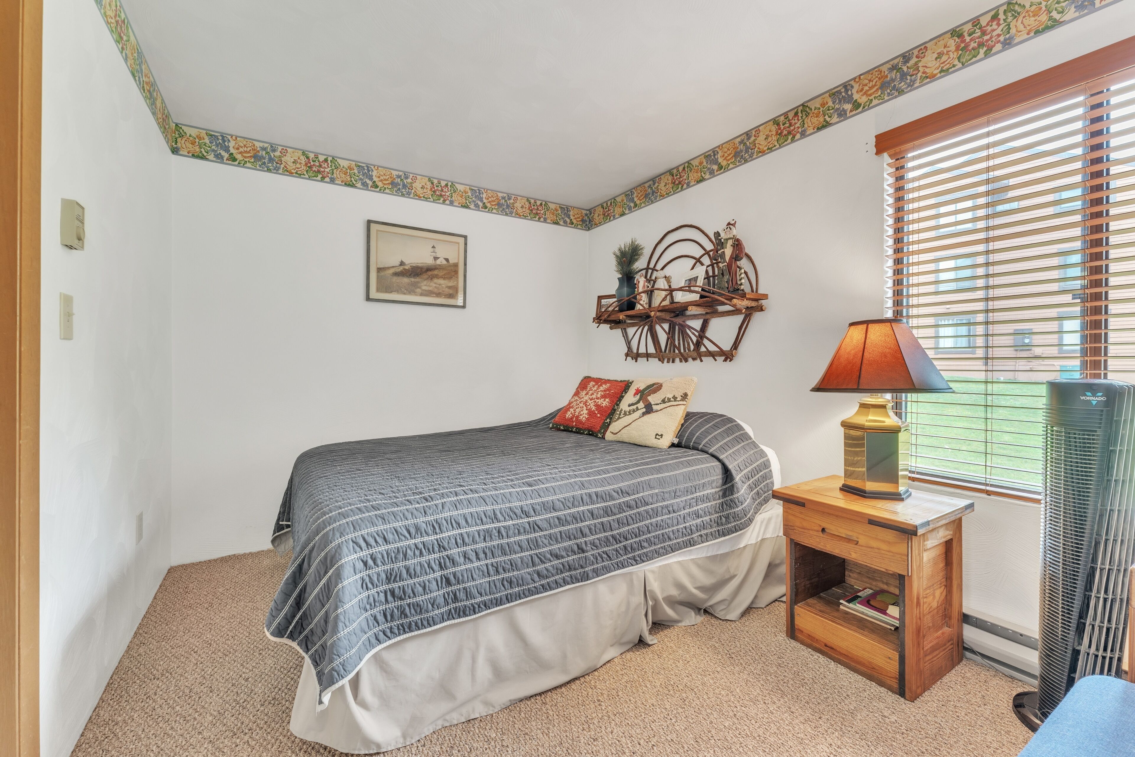 A bedroom with a bed, a nightstand, a lamp, a radiator, a window with blinds, and a wall with a floral border.