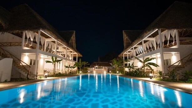 A swimming pool with a clear blue water surface, surrounded by buildings with thatched roofs and white walls. - Rafiki Tamu Residential Resort (Watamu)