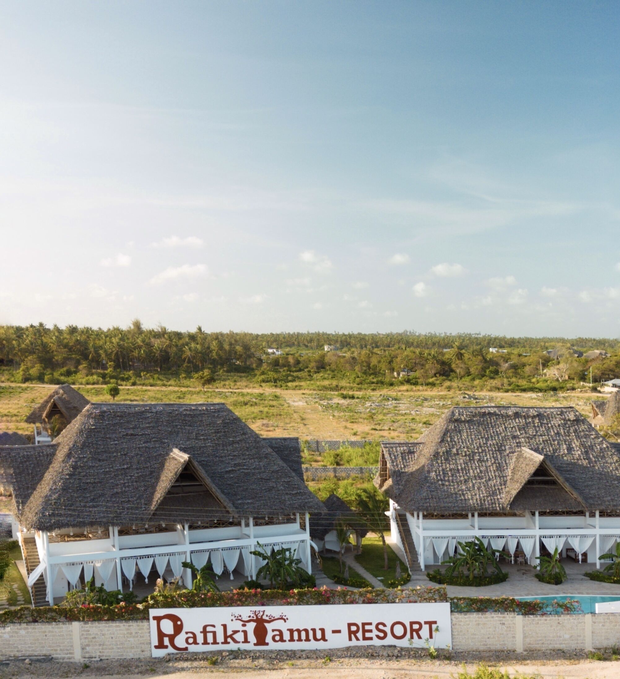A resort with thatched-roof buildings, a sign reading "Rafiki Tamu - Resort", and a view of the surrounding landscape.