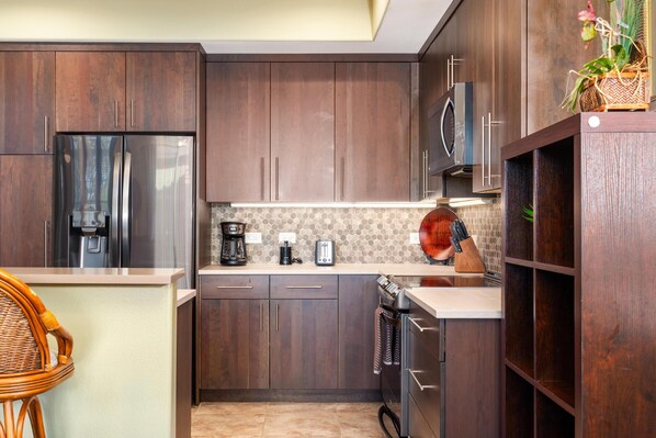 A modern kitchen with dark wood cabinets, stainless steel appliances, and a herringbone tile backsplash. - Big Island Waikoloa Colony Villas 1206 2 Bedroom Condo (Waikoloa)