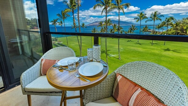 A balcony with wicker furniture, a table set for two, and a view of a golf course and ocean. - Kihei Surfside 412 1 Bedroom Condo (Kihei)