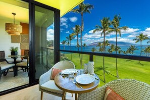 A balcony with a table set for dining, overlooking a lawn with palm trees and a view of the ocean. - Kihei Surfside 412 1 Bedroom Condo (Kihei)
