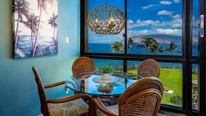 A dining area with a glass table, wicker chairs, and a view of the ocean through large windows. - Kihei Surfside 506 1 Bedroom Condo (Kihei)